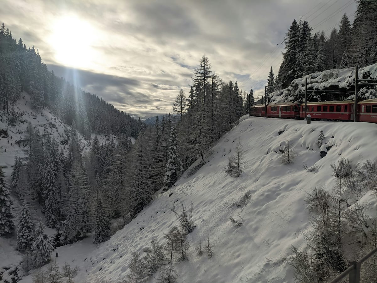 A red Bernina train runs along a snowy hillside above a forested valley on a winter day.