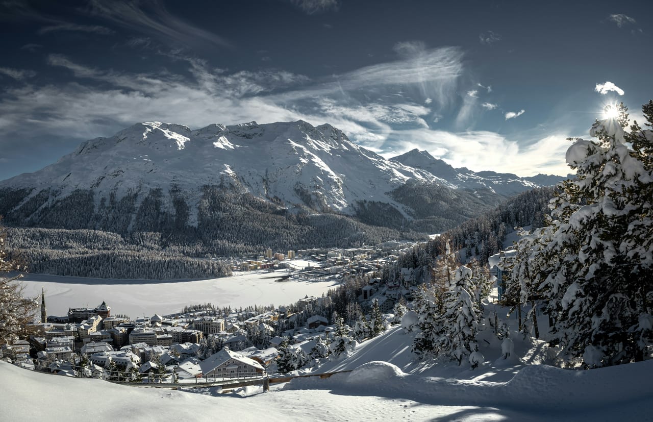 St. Moritz in winter: snow-covered rooftops above the frozen lake, with forested slopes and snowy peaks beyond.